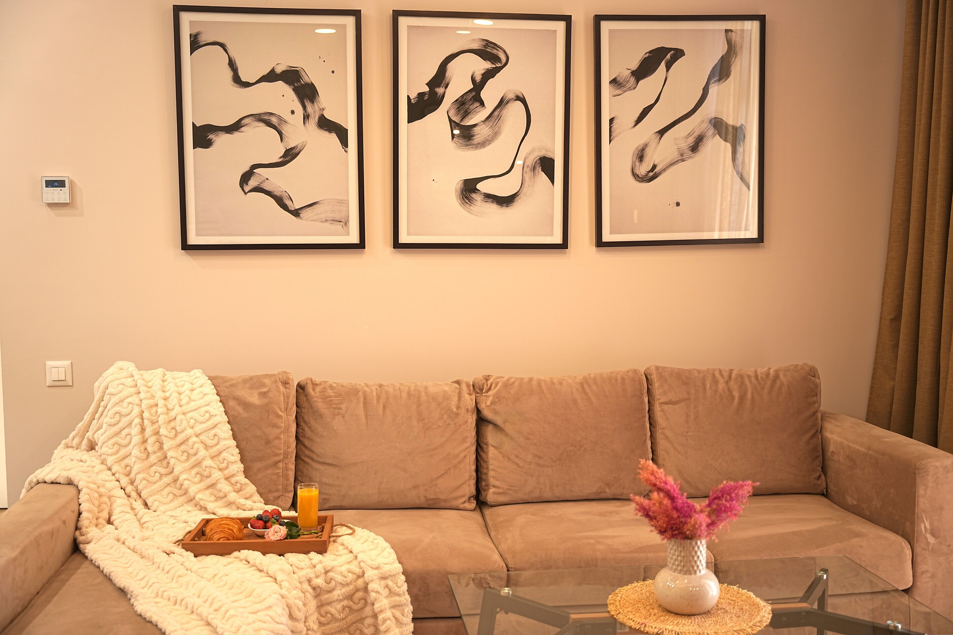 Un salon avec un canapé beige, une table basse en verre et trois tableaux abstraits accrochés au mur.