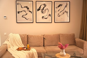 A living room with a beige sofa, a glass coffee table, and three abstract paintings on the wall.