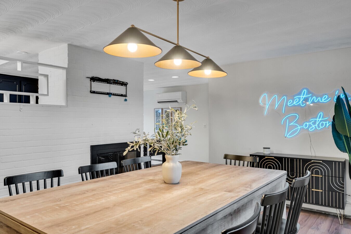 A modern dining area with a wooden table, black chairs, and a neon sign reading "Meet me in Boston".