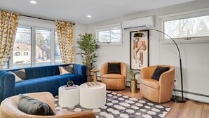 A modern living room with a blue sofa, two armchairs, a coffee table, and a floor lamp. There is a large window with curtains, a potted plant, and a wall-mounted air conditioning unit. - Luxury Boston Ranch –Sleeps 8|Pet+|Heated pool (Framingham)