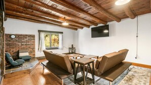 Une pièce chaleureuse avec une cheminée en briques, des poutres apparentes en bois au plafond, un téléviseur fixé au mur et un coin repas avec une table et des bancs.
