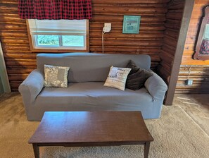 A log cabin interior with a sofa, two pillows, a small table, a window with blinds, a wall-mounted power outlet, and a framed picture. - Catalina - Very Cute, Comfy, and Traditional!  Includes Window A/C Unit and 2 Kayaks!  Dog-friendly as well! (Oscoda)