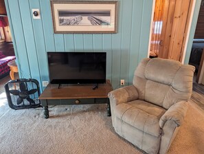 A living room with a television on a wooden stand, a beige armchair, and a framed picture on the wall. - Catalina - Very Cute, Comfy, and Traditional!  Includes Window A/C Unit and 2 Kayaks!  Dog-friendly as well! (Oscoda)
