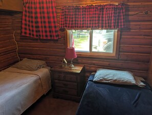 A cabin-style room with two beds, a window with curtains, a lamp, and a small wooden dresser. - Catalina - Very Cute, Comfy, and Traditional! Includes Window A/C Unit and 2 Kayaks! Dog-friendly as well! (Oscoda)