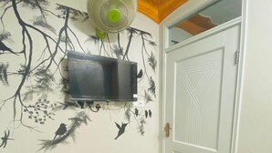 Une pièce avec un sol carrelé, un téléviseur fixé au mur, un ventilateur sur pied, une porte ornée de motifs géométriques et une poubelle.