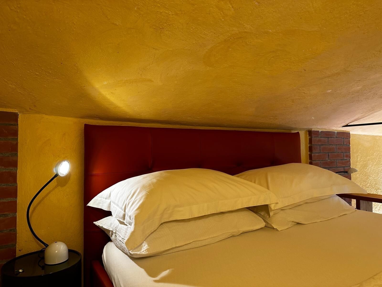 Quarto com cabeceira vermelha, abajur e parede de tijolos.