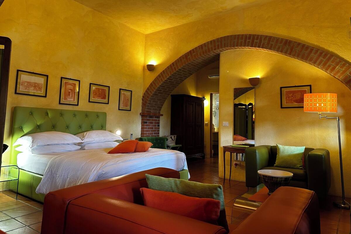 A hotel room with a large bed, green upholstered headboard, and a brick archway.