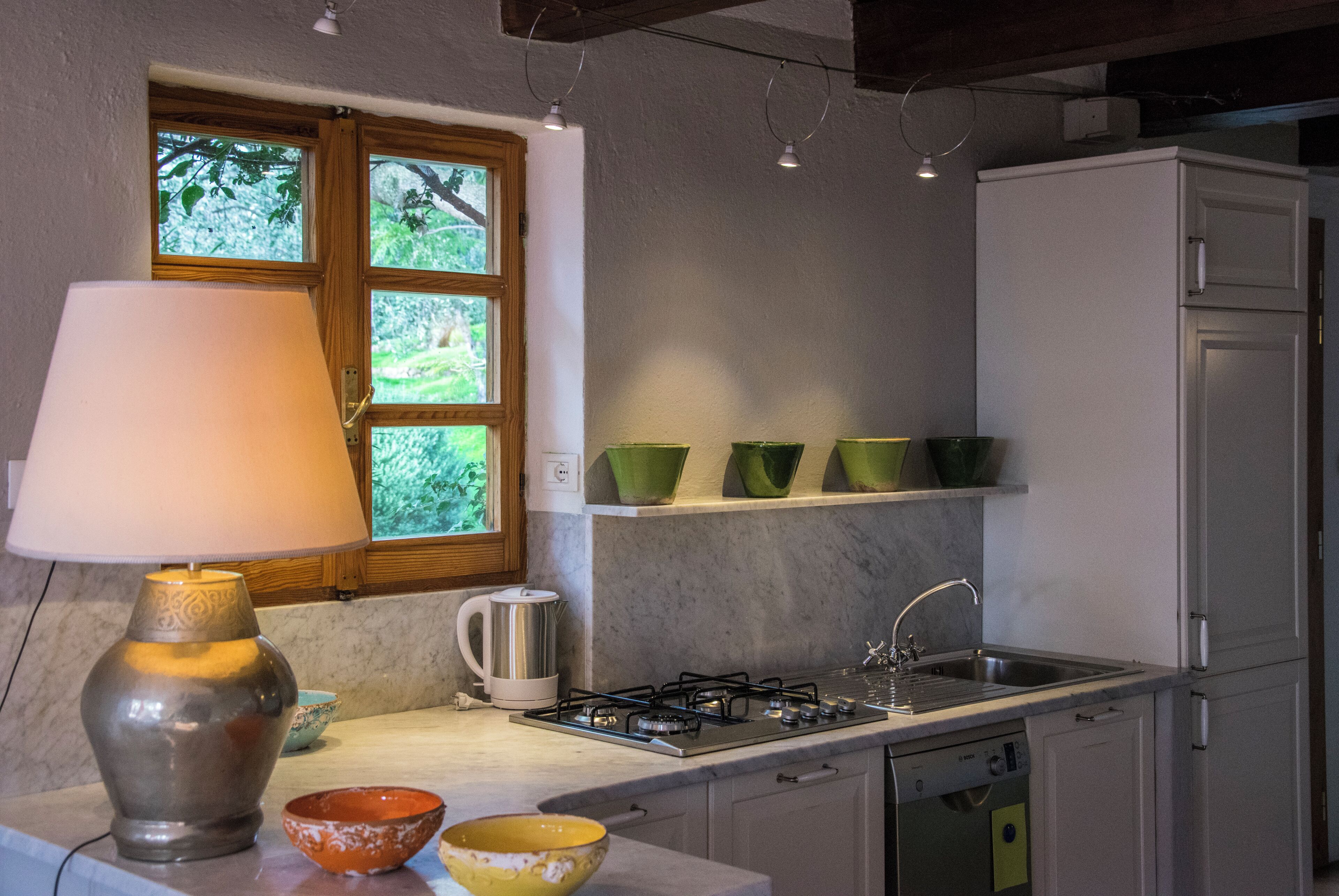 Une cuisine équipée d’une cuisinière, d’un évier et de placards. Il y a une fenêtre donnant sur des arbres et une lampe sur le plan de travail.