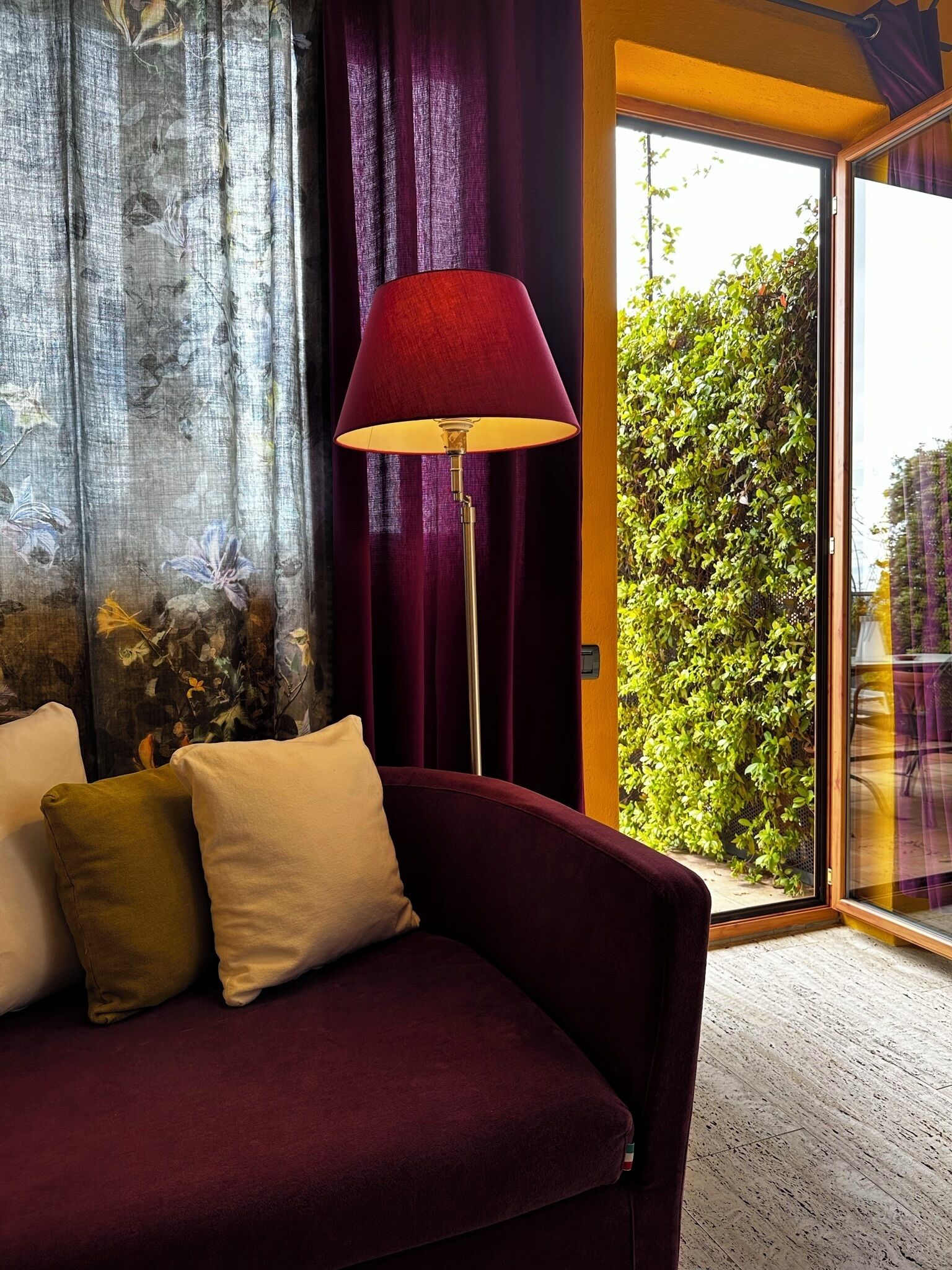 A room with a dark red sofa, a floor lamp, and a view of greenery through a glass door.