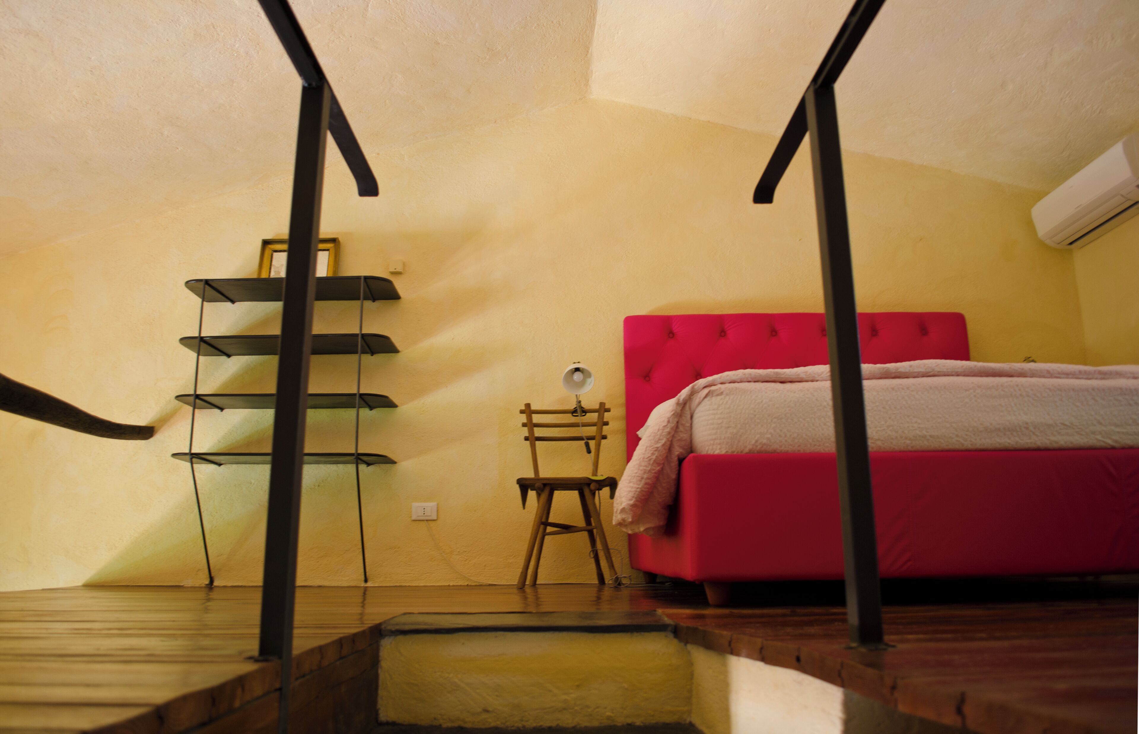 A bedroom with a red bed, a wooden chair, a small table with a lamp, and a shelf unit.
