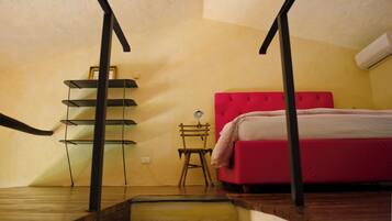 A bedroom with a red bed, a wooden chair, a small table with a lamp, and a shelf unit.