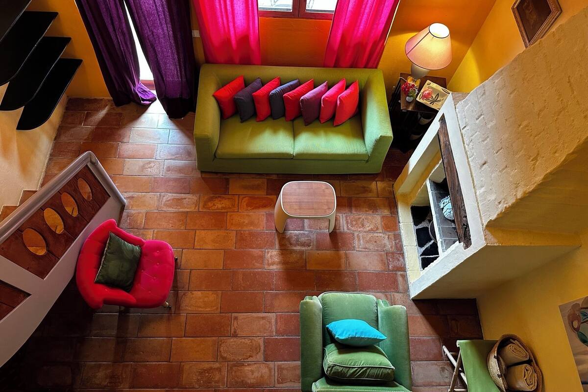 A cozy living room with a green sofa, red and black cushions, a red chair, and a green armchair. There is a small table and a lamp on a side table. The room has a brick floor and a window with curtains.