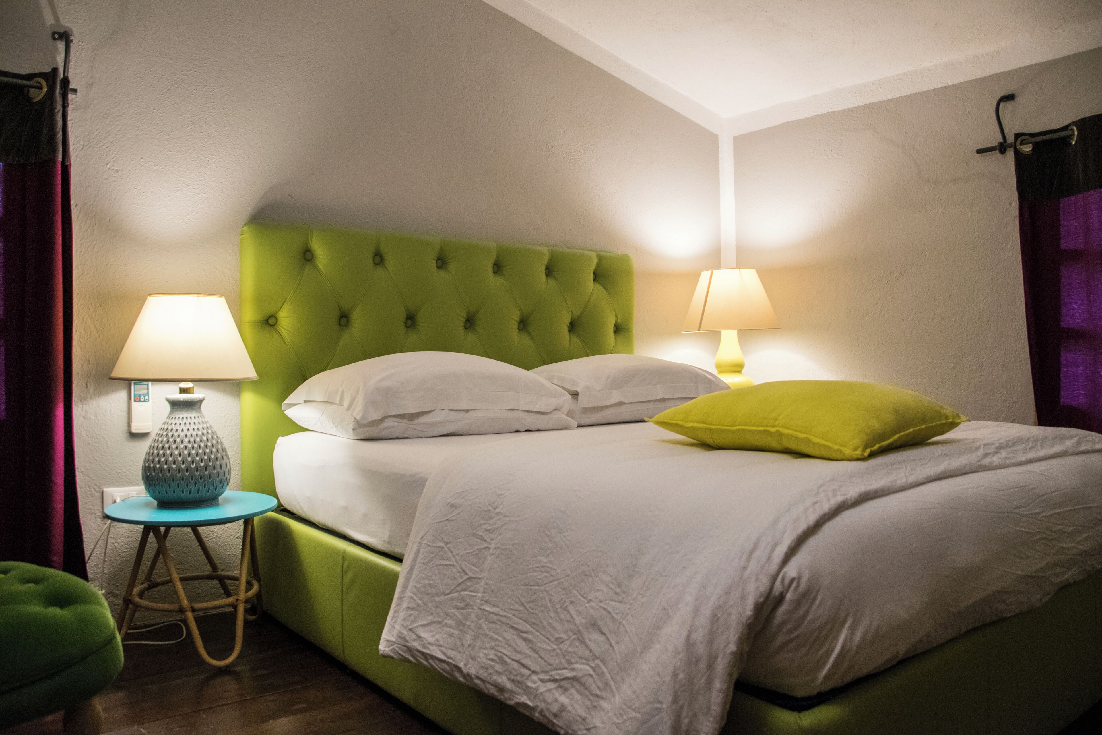 Ein ordentlich bezogenes Bett mit weißen Leinen und einem grünen Kopfteil, zwei Nachttische mit Lampen sowie ein kleiner runder Tisch ebenfalls mit einer Lampe.