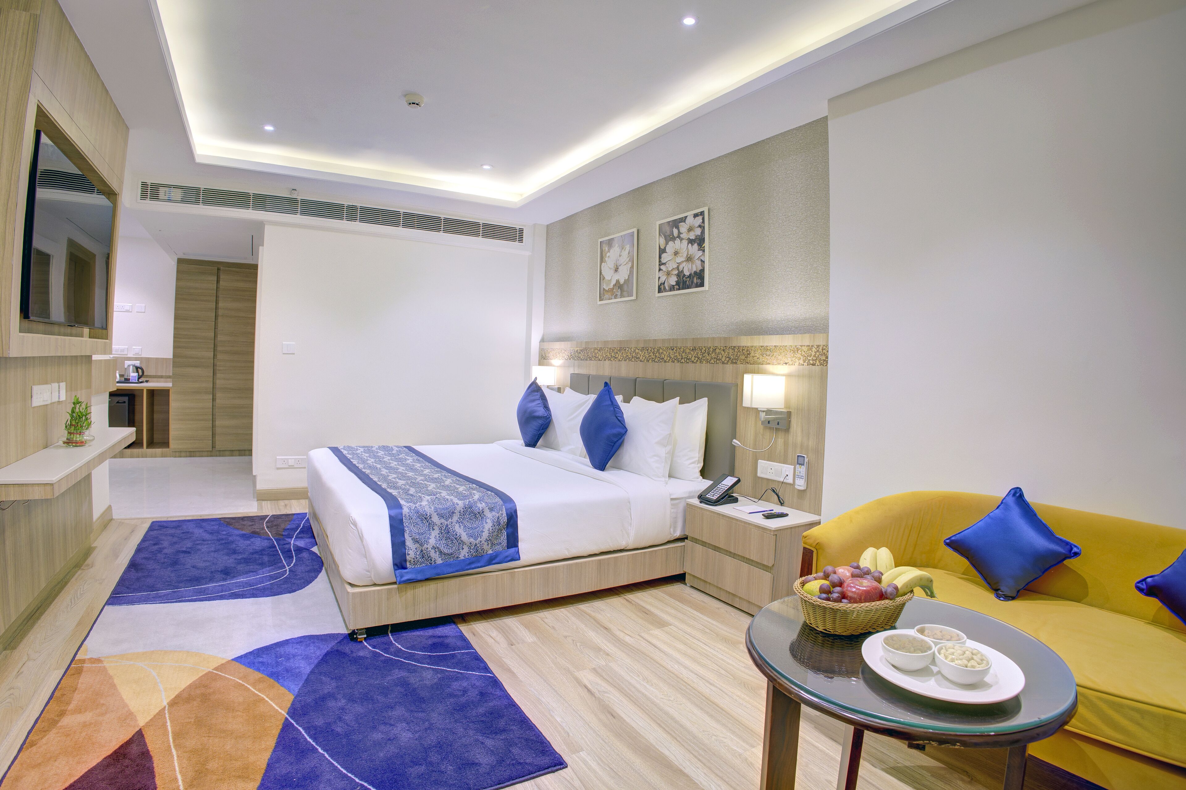A modern hotel room with a bed, a sofa, a dining table with a fruit basket, and a small kitchen area.
