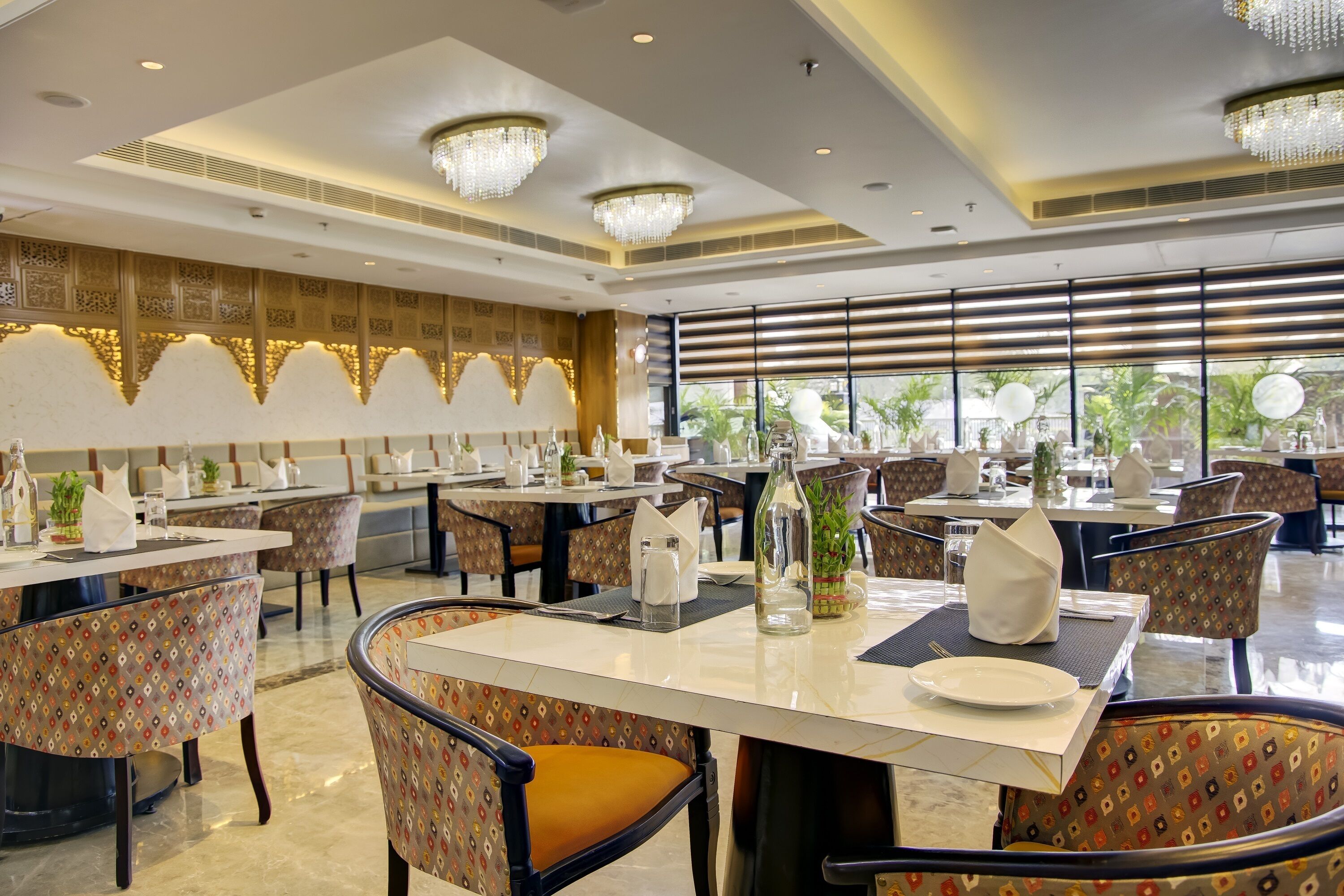 A spacious dining area with multiple tables and chairs, chandeliers, and large windows with views of greenery.