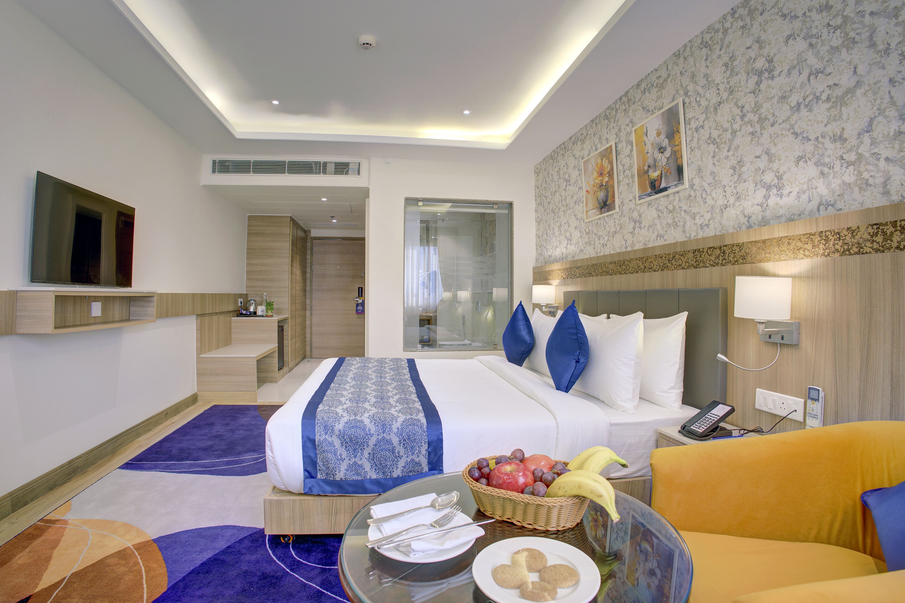 A hotel room with a bed, a small table with breakfast items, a chair, a TV, and a bathroom visible through a glass door.