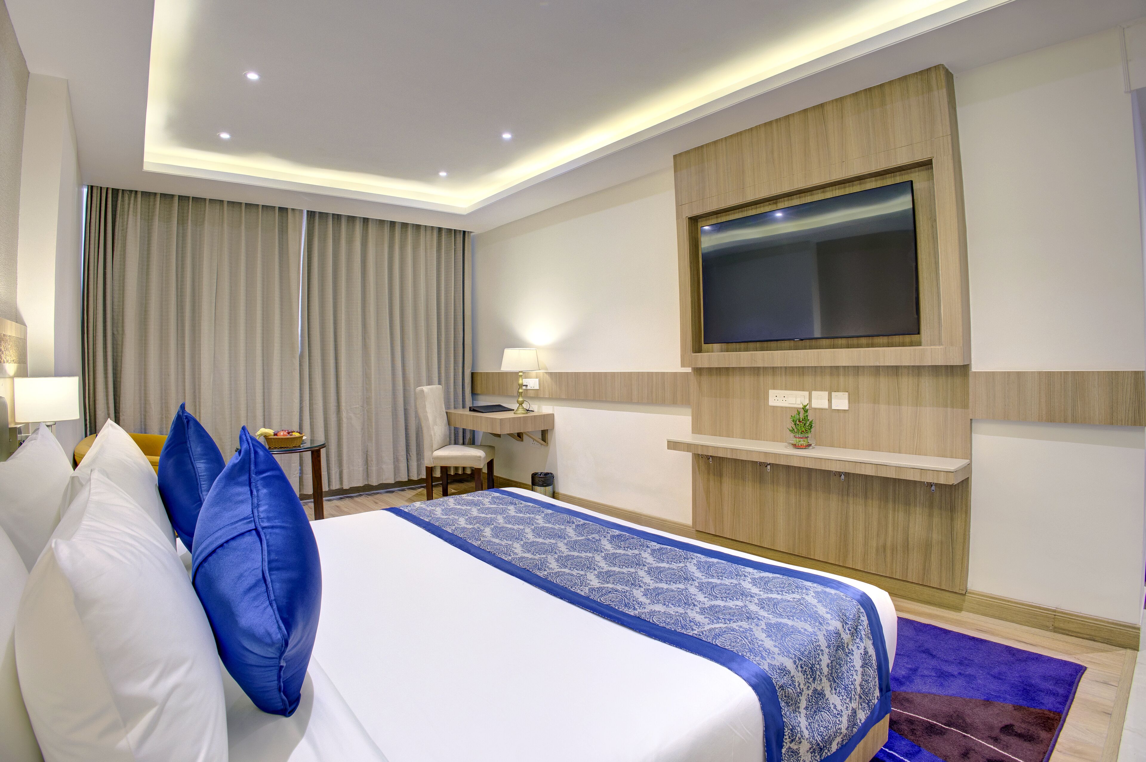 Habitación de hotel con una cama grande, un escritorio, una silla, un televisor montado en la pared y una ventana con cortinas.