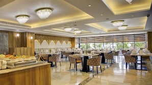 Un vaste espace dédié à la salle à manger, avec de nombreuses tables et chaises rondes, des lustres et un buffet proposant une variété de plats.