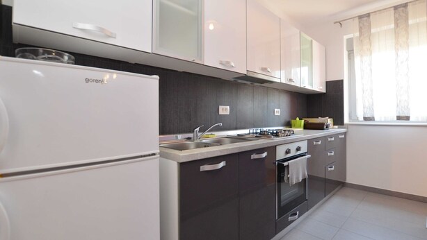 A modern kitchen with a white refrigerator, dark cabinets, a stainless steel sink, and a window with sheer curtains. - Quiet and Compact Apartment With Balcony (Rovinj)