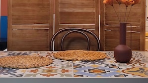 Een eetruimte met een tafel gedekt voor twee personen, een geweven placemat, een vaas met bloemen, en een houten scheiding met een ronde lamp erboven.