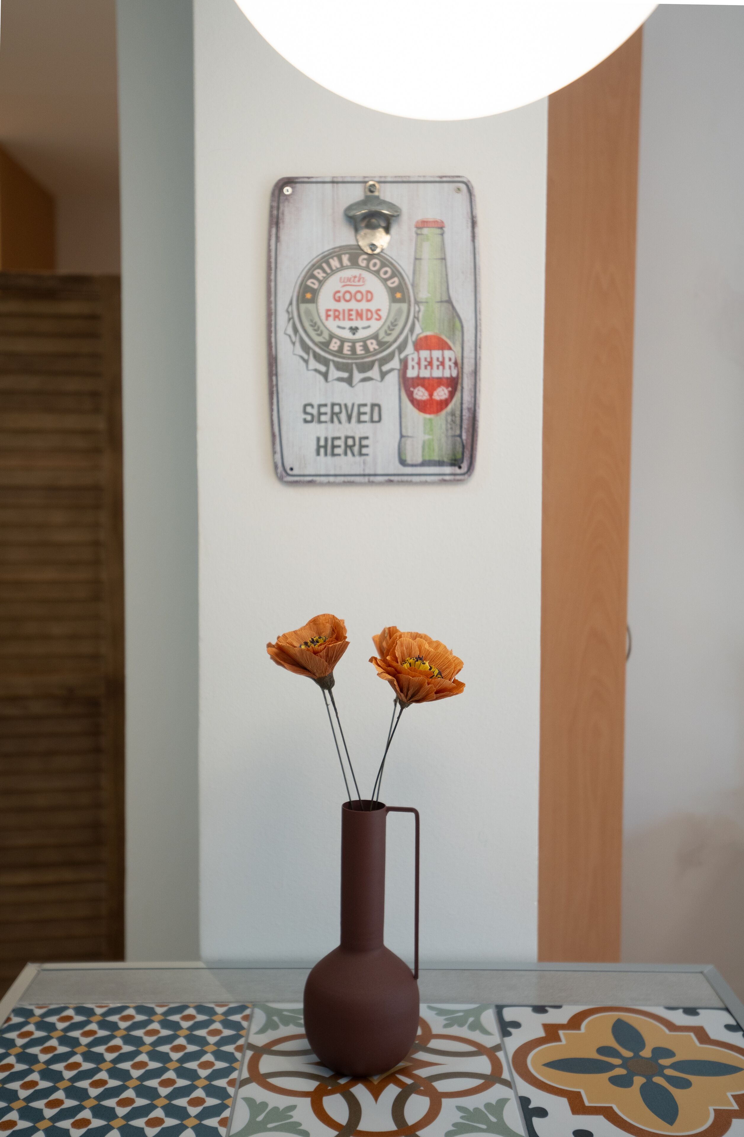 A wall-mounted sign with a beer bottle illustration and the text "DRINK GOOD with GOOD FRIENDS BEER SERVED HERE." Below the sign, there is a vase with two orange flowers on a table with patterned tiles.