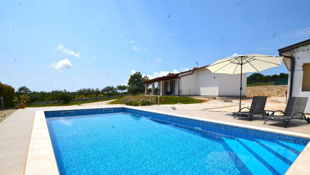 Piscina rectangular con azulejos azules, rodeada por un solárium de concreto. Hay dos sillas de descanso y una sombrilla bajo una pérgola.