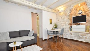 Un soggiorno moderno con un divano grigio, un tavolino rotondo e un tavolo da pranzo con sedie. Si nota una parete in pietra con una TV a parete e una sveglia.