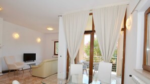 A modern living room with a glass table, white furniture, a flat-screen TV, and a balcony with white curtains. - Elegant and Brigth Corner Apartments (Rovinj)