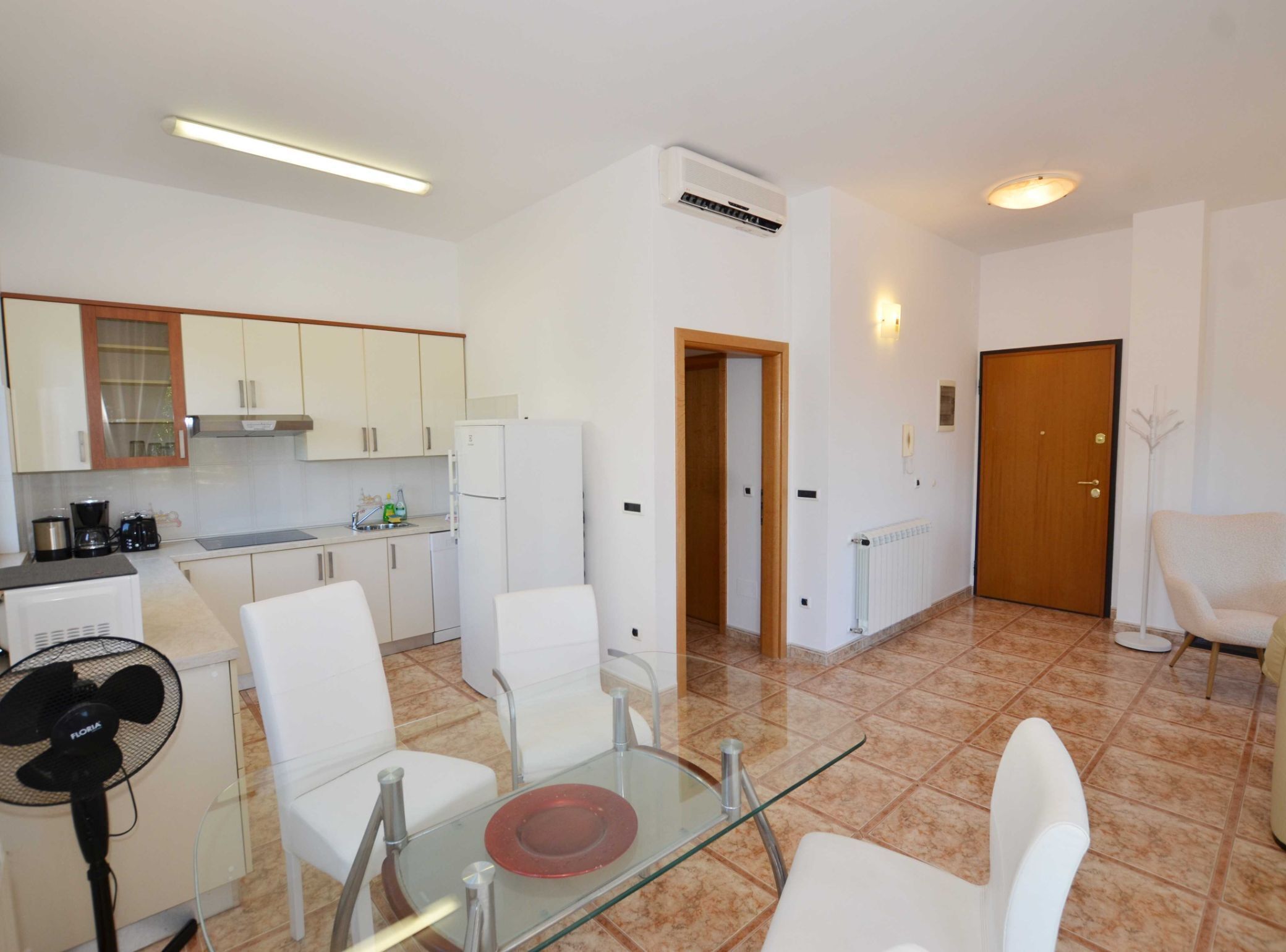 Una cocina moderna con armarios blancos, un refrigerador y una cafetera. Hay un comedor con una mesa de vidrio y sillas blancas.