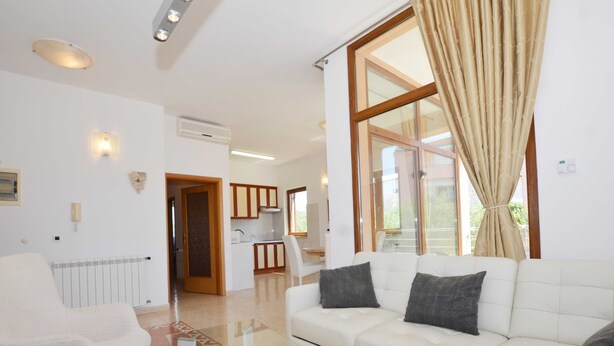 A modern living room with a white sofa, a glass coffee table, and a white armchair. There is a kitchen in the background with wooden cabinets and a window with curtains. - Elegant and Brigth Corner Apartments (Rovinj)