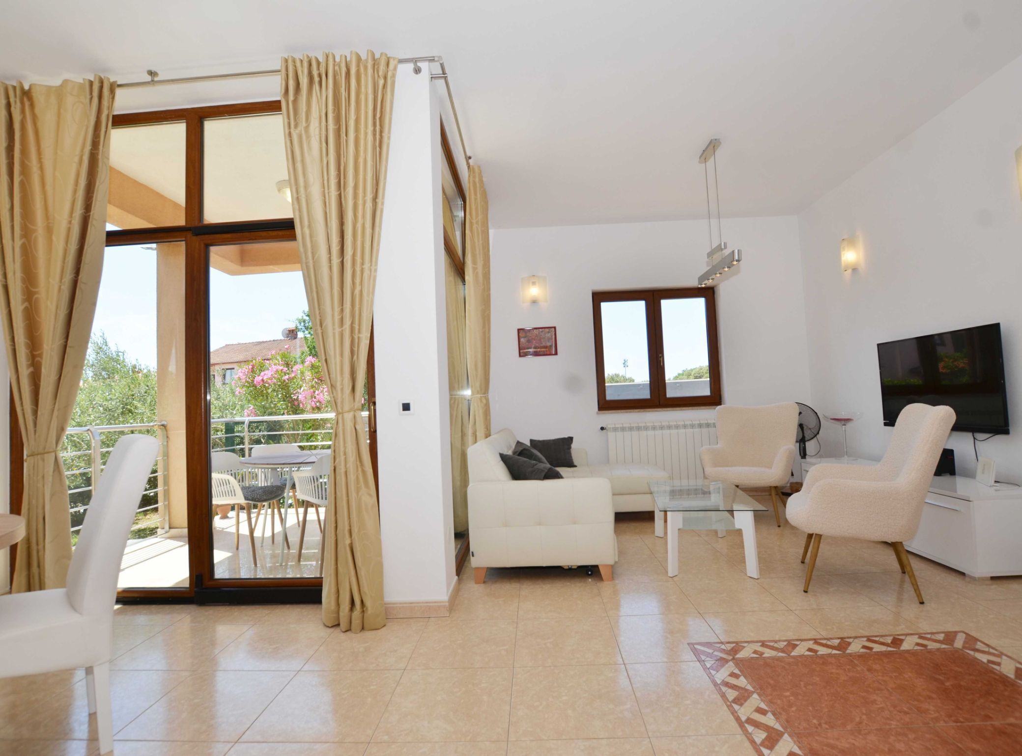 A spacious living room with a sliding glass door leading to a balcony, a flat-screen TV mounted on the wall, and a dining area with a table and chairs.