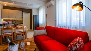 A modern living room with a red sofa, a dining area with wooden chairs, a kitchen with white cabinets, and a large window with sheer curtains. - Modern and Stylish Rovinj Apartments With Terrace (Rovinj)