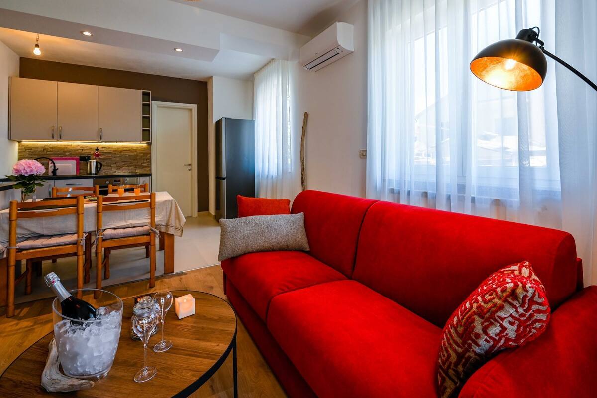 Un salon moderne avec un canapé rouge, un coin repas avec des chaises en bois, une cuisine avec des meubles blancs et une grande fenêtre ornée de rideaux légers.