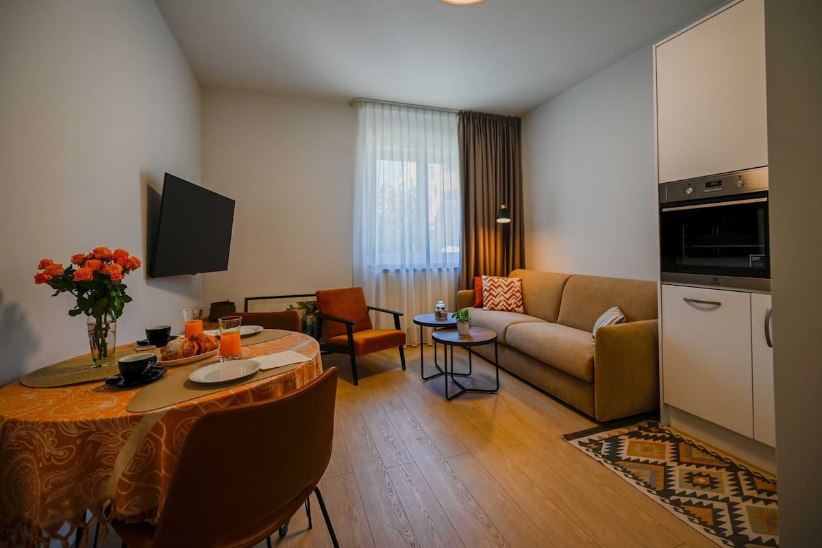 Un salon moderne comprenant un canapé, une table à manger pour deux personnes, un téléviseur fixé au mur et un coin cuisine avec un four.