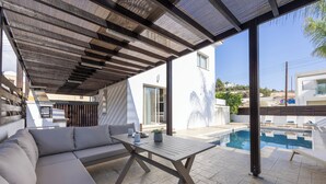 A covered outdoor seating area with a pool and a modern building in the background.