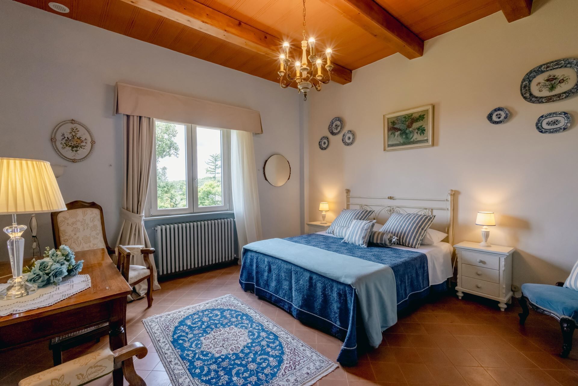 A bedroom with a bed, bedside tables, a desk, a chair, a radiator, a window, and a chandelier.