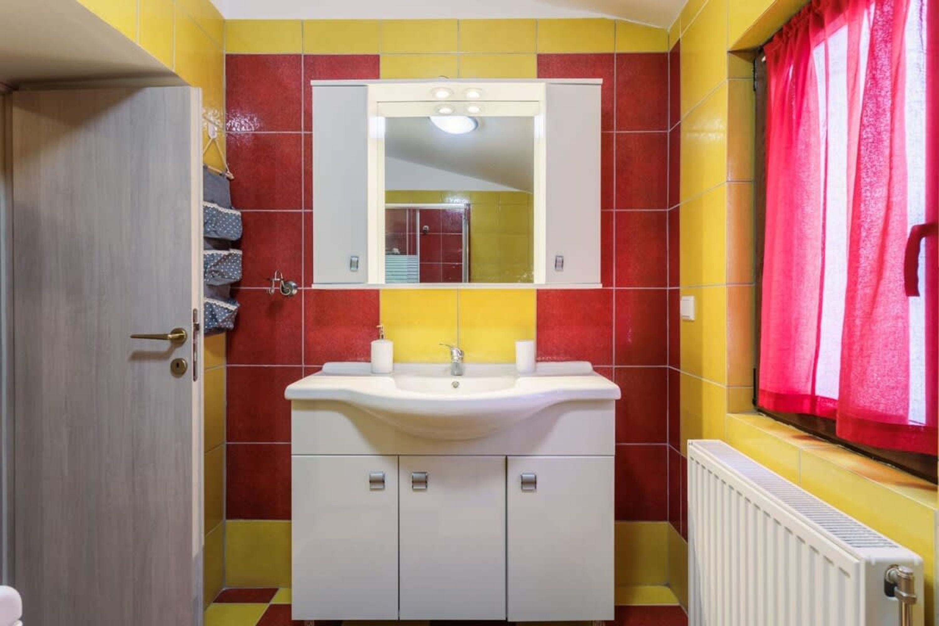 A bathroom with a white sink, mirror, and red and yellow tiled walls. There are towels hanging on the wall and a window with red curtains.