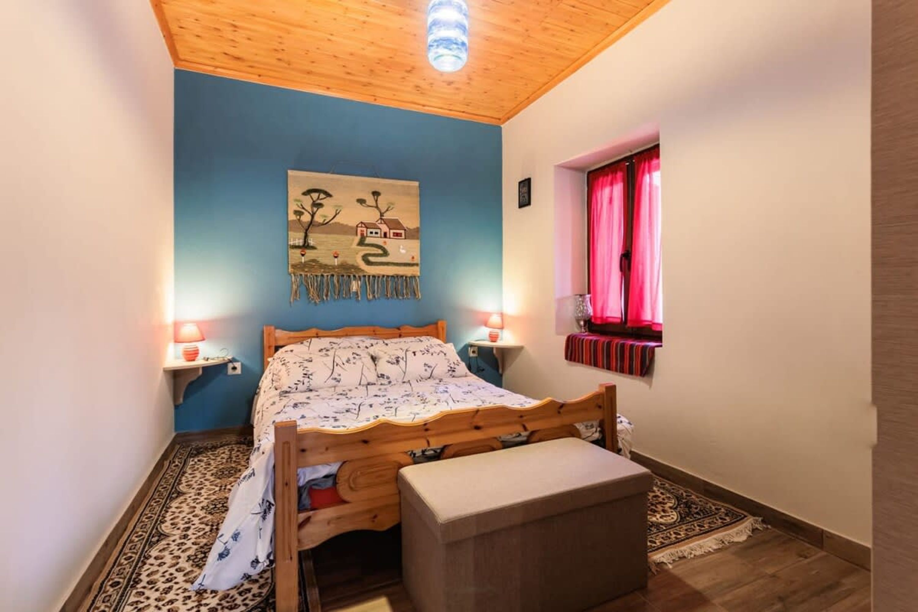 Camera da letto con un letto in legno, una parete azzurra, un quadro, due lampade ai lati del letto e una finestra con tende rosse.