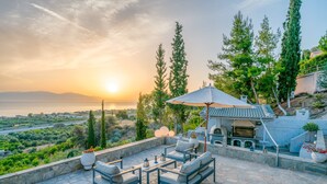 An outdoor patio with stone flooring, seating, and a barbecue area, overlooking a scenic landscape with trees and a view of the sea.