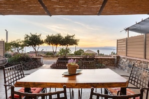 Une terrasse extérieure avec une table et des chaises en bois, un panier en osier rempli de fleurs et une vue sur la mer et les montagnes.