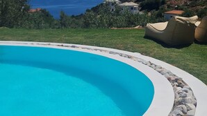 A clear blue swimming pool with a white edge, surrounded by a stone border. In the background, there are houses and a view of the sea. - Estia Seaview Room - Ariadne Skopelos Getaway (Skopelos)