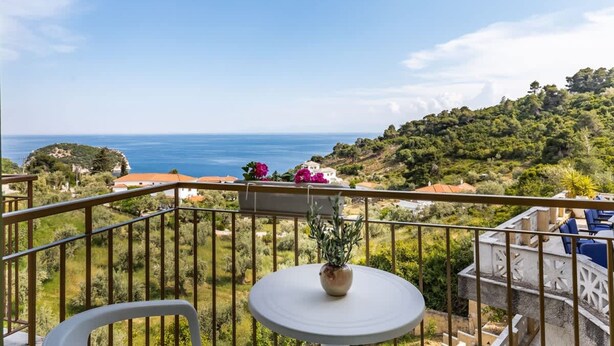 A balcony with a white table and chairs, overlooking a coastal landscape with houses and greenery. - Estia Seaview Room - Ariadne Skopelos Getaway (Skopelos)