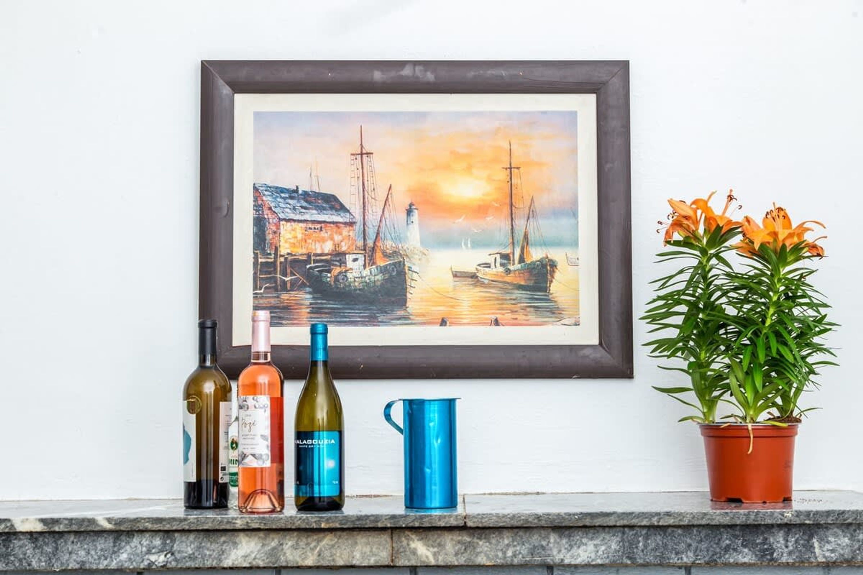 Un tableau encadré représentant un port avec des bateaux et un phare, une bouteille de vin, un verre à vin et une plante en pot aux fleurs oranges, le tout posé sur un rebord en marbre.