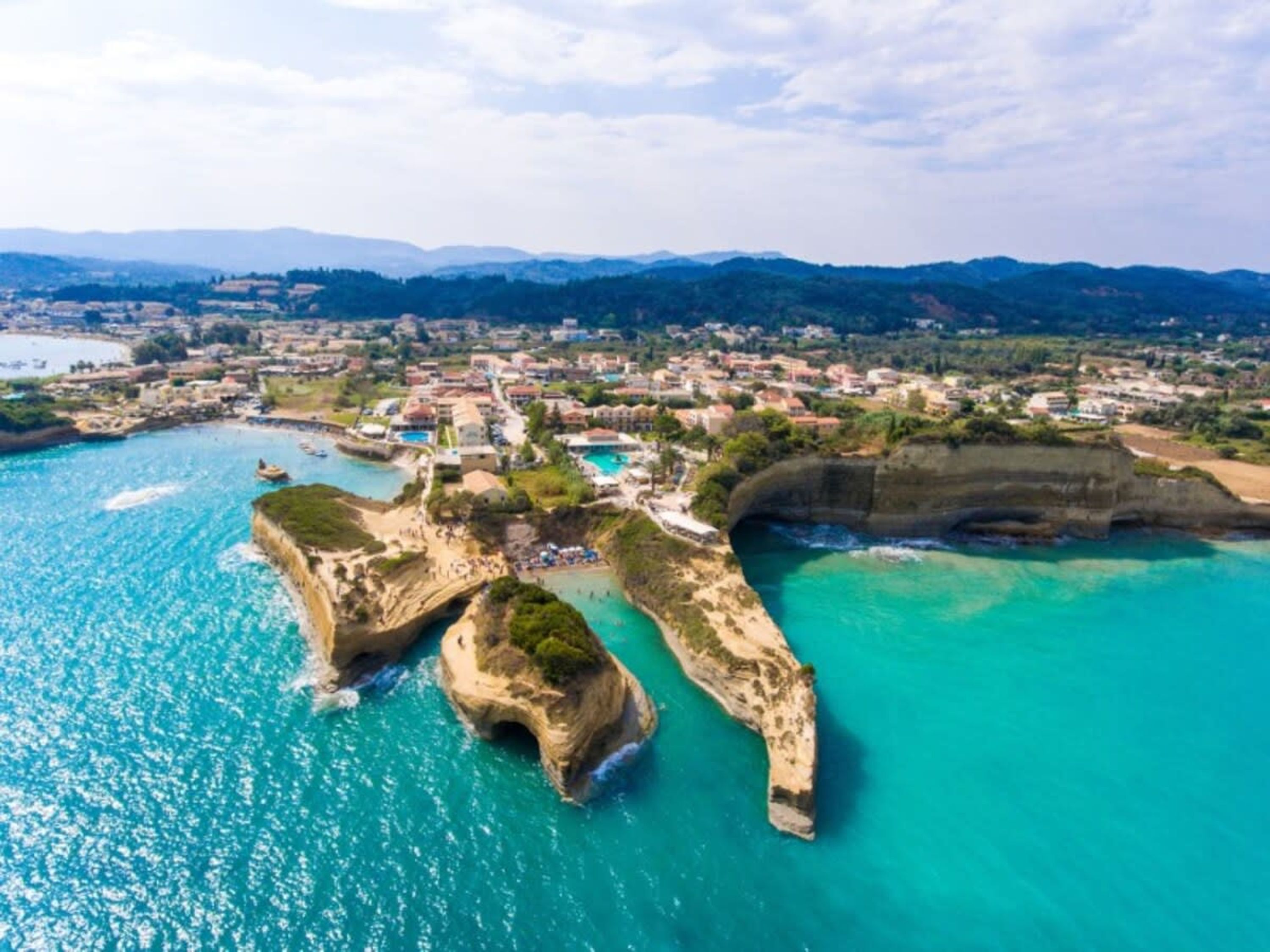Vista aérea de un pueblo costero con aguas turquesas cristalinas, formaciones rocosas y una combinación de edificios residenciales y comerciales.
