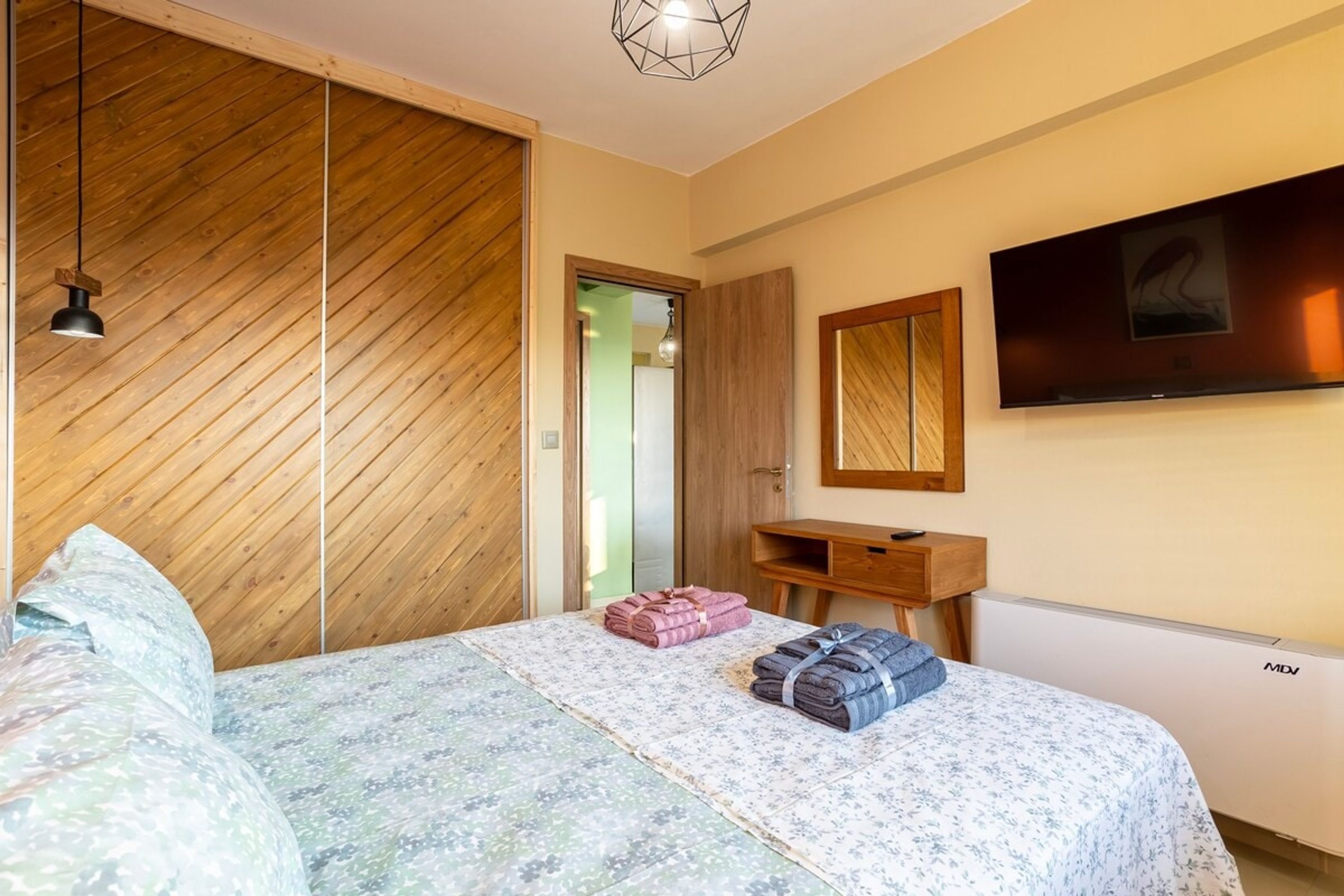 A bedroom with a wooden slat ceiling, a bed with a floral bedspread, a wooden desk, a mirror, and a mounted TV.