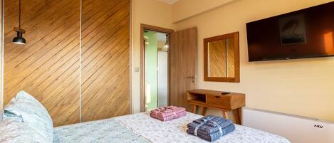 A bedroom with a wooden slat ceiling, a bed with a floral bedspread, a wooden desk, a mirror, and a mounted TV.