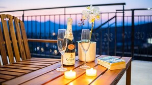 A wooden table with a bottle of champagne, two glasses, a candle, and a book on a balcony with a railing and a view of the mountains at dusk. - Martinia Pool Escape - Tragos Panoramic Views (Kalamata)