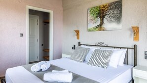 A neatly made bed with white linens and patterned pillows, a painting of a tree on the wall, and a wooden bench against the wall. - Persephone Seaview Room - Ariadne Skopelos Getaway (Skopelos)
