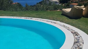 A swimming pool with a curved edge, surrounded by a stone border. In the background, there are houses and a view of the sea. - Persephone Seaview Room - Ariadne Skopelos Getaway (Skopelos)
