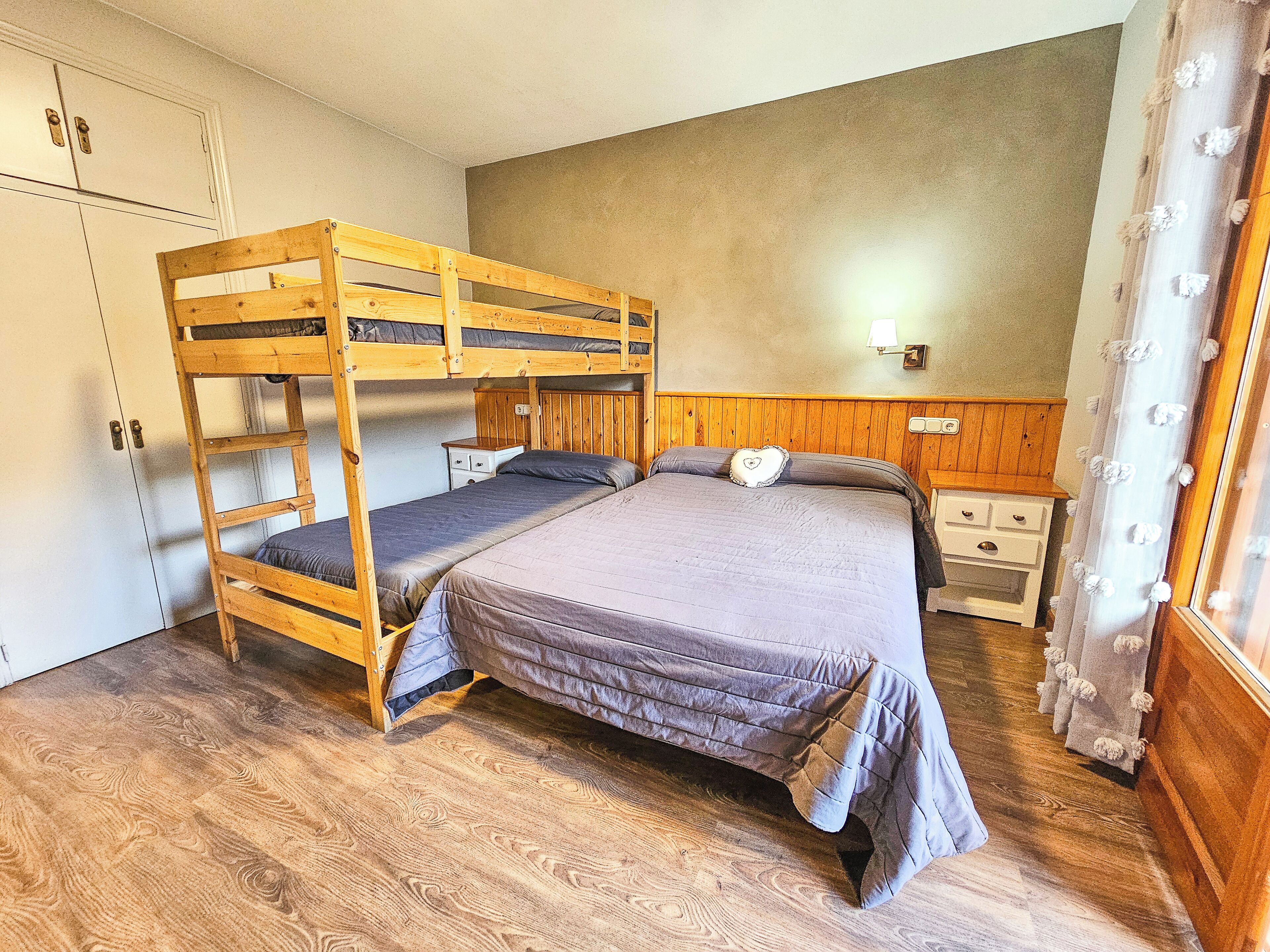 A bunk bed room with a wooden bunk bed, a single bed, a nightstand, a dresser, and a wall-mounted lamp.